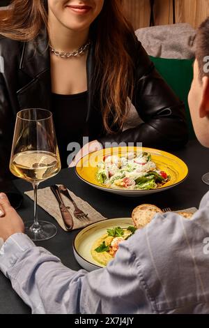 Junges Paar genießt ein romantisches leichtes Abendessen bei einem Glas Weißwein mit Caesar Salat und Hummus mit gebratenen Garnelen, zugeschnittenem Shot Stockfoto