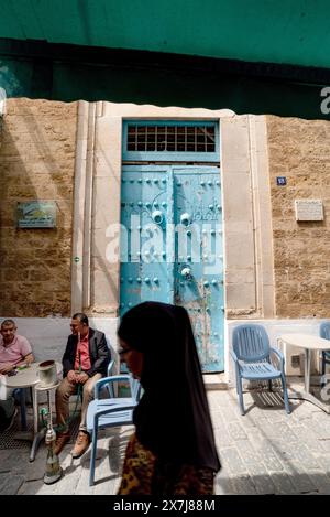 Tunis, Tunesien. 13. Mai 2024 eine alte blaue Holztür mit tunesischen Männern, die Shisha-Wasserpfeifen rauchen, in der geschäftigen Medina von Tunis, einem UNESCO-Weltheritag Stockfoto