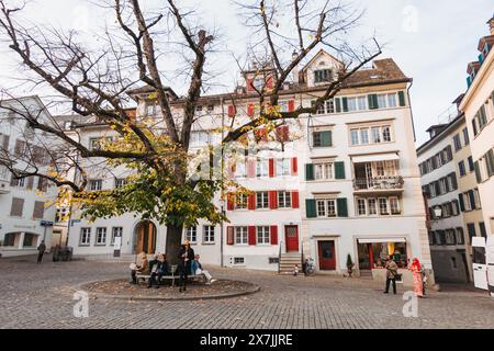 Die Menschen sitzen im Herbst unter einem großen Laubbaum auf einem Zürcher Stadtplatz, umgeben von historischen Wohnhäusern mit bunten Fensterläden Stockfoto