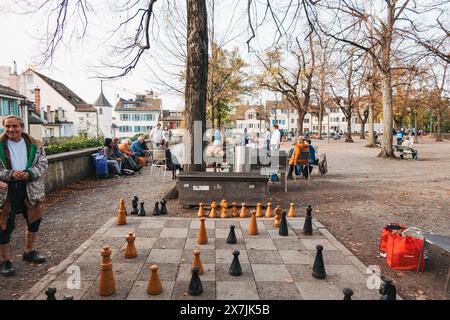 Ein Mann steht neben einem großen Schachbrett im Park Lindenhof, Zürich, wo sich die Menschen auf Bänken und unter Herbstbäumen entspannen Stockfoto