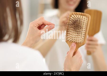 Frau, die ihr verlorenes Haar von der Bürste in der Nähe des Spiegels in Innenräumen nimmt, Nahaufnahme. Alopezie-Problem Stockfoto