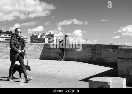 Sonniger Tag auf der Löwenbrücke: Eine Frau in warmem Outfit und Sonnenbrille geht zügig mit ihrem Hund spazieren, ein älterer Mann liest ein Buch mit unbeschwerter Haltung. Stockfoto