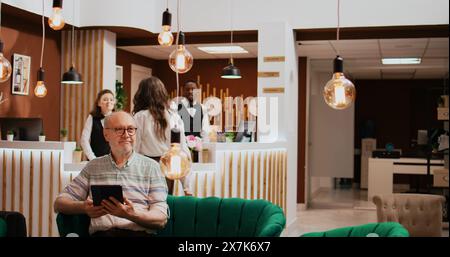 Ein älterer Mann überprüft die Verbindung des Tablets im Hotel und entspannt sich im Loungebereich, bevor er in den Ruhestand geht. Auf Online-Websites finden Sie Freizeiteinrichtungen und entspannende Aktivitäten. Stockfoto