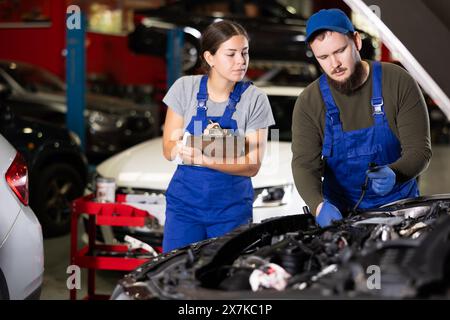 Eine Frau mit Dokumenten sieht einen Mechaniker an, der das Auto unter der Motorhaube repariert Stockfoto