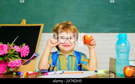 Gesundes Frühstück für Schüler. Lächelnder süßer Junge mit Apfel während der Mittagspause im Klassenzimmer. Kleiner Schuljunge in Brille am Schreibtisch davor Stockfoto