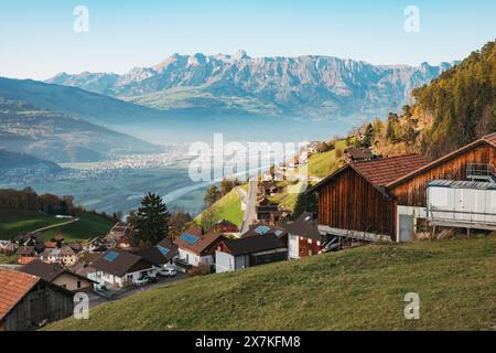 Ein malerisches Dorf mit Holzhäusern auf einem Hügel in Liechtenstein, mit Blick auf ein Tal mit einer gewundenen Straße und einer Stadt in der Ferne Stockfoto