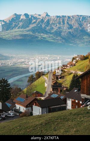 Ein malerisches Dorf mit Holzhäusern auf einem Hügel in Liechtenstein, mit Blick auf ein Tal mit einer gewundenen Straße und einer Stadt in der Ferne Stockfoto