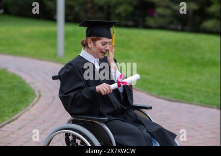 Eine weiße Frau im Rollstuhl hält ihr Diplom und weint vor Freude draußen. Stockfoto