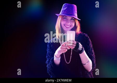 Rothaarige junge Frau mit lila Paillettenhut und Sonnenbrille benutzt ihr Handy und lächelt selbstbewusst. Sie trägt ein stylisches schwarzes Oberteil und Jeans und ist surr Stockfoto