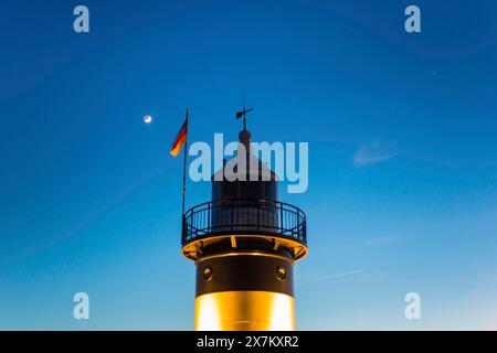 Der schwarz-weiße Leuchtturm „kleine Preusse“ steht kurz nach Sonnenuntergang nachts vor dem blauen Himmel, dem wachsenden Mond, beleuchtet Stockfoto