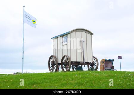 Alter Holzwagen, Badewagen mit Zugstange und großen Holzrädern auf grüner Wiese mit Fahne daneben, im Hintergrund Strandstuhl, bewölkt Stockfoto