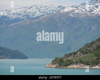 Ein ruhiger Seeufer mit Bergen und bewaldeten Hügeln, unter einem klaren blauen Himmel mit Wolken, grünlich glitzerndem Wasser in einem Fjord mit schneebedeckten Bergen Stockfoto