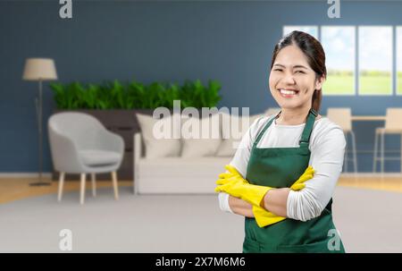 Die asiatische Haushälterin trägt gelbe Gummihandschuhe und eine grüne Schürze, die im Wohnzimmer lächelt Stockfoto