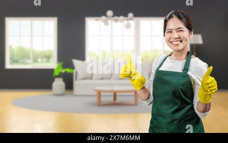 Die asiatische Haushälterin trägt gelbe Gummihandschuhe und eine grüne Schürze, die im Wohnzimmer lächelt Stockfoto