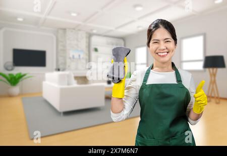 Die asiatische Haushälterin trägt gelbe Gummihandschuhe und eine grüne Schürze, die im Wohnzimmer lächelt Stockfoto