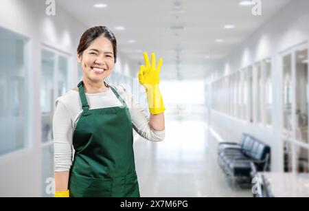 Asiatische Haushälterin trägt gelbe Gummihandschuhe und grüne Schürze im Büro Stockfoto