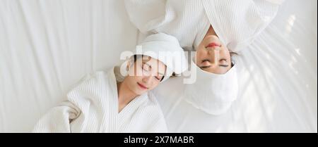 Zwei asiatische Frauen liegen in weißen Bademänteln und Handtüchern um den Kopf gewickelt auf einem Bett. Draufsicht Stockfoto