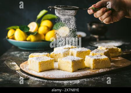 Seitlicher Aktionsfoto von weiblicher Hand, die Puderzucker über Zitronenkuchen-Quadrate streut, Schüssel Zitronen in BG Stockfoto