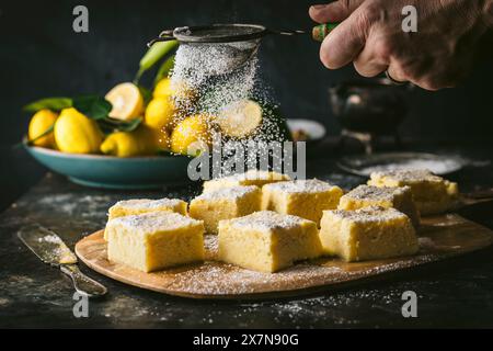 Seitliche Winkelaktion von männlicher Hand, die Puderzucker über Zitronenkuchen-Quadrate streut, Schüssel Zitrone in BG Stockfoto