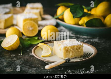 Seitenwinkel der quadratischen Zitronenkuchenscheibe auf dem Dessertteller, mit Zitronengarnitur und frischen Zitronen im Hintergrund Stockfoto