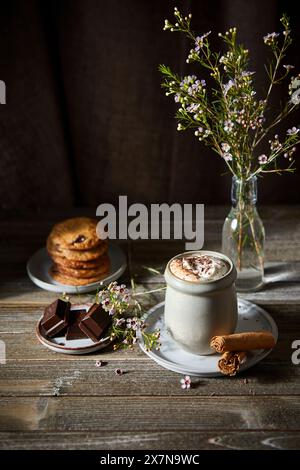 Mexikanische heiße Schokolade mit Schlagsahne auf dunklem Holzhintergrund mit Blumen, Keksen und Zimtstangen. Stockfoto