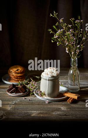 Mexikanische heiße Schokolade mit Schlagsahne auf dunklem Holzhintergrund mit Blumen, Keksen und Zimtstangen. Stockfoto