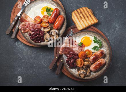 Traditionelles englisches Frühstück mit Spiegeleiern, Würstchen, Bohnen, Pilzen, gegrillten Tomaten, Speck und Toast auf grauen Tellern Stockfoto