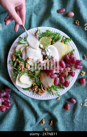 Salat im Waldorfstil mit Birnen, Walnüssen, Sellerie, Trauben und Baby-Raketenblättern. Weibliche Hand, die einen Verband hinzufügt. Stockfoto