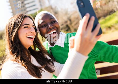 Zwei junge Freundinnen lachen glücklich und haben Spaß, Selfie-Fotos mit Handy in einem Stadtpark zu machen, Konzept der Vielfalt und modernen Lebensstil Stockfoto
