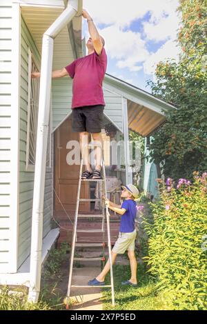 Der Sohn hält die Trittleiter, auf der sein Vater an sonnigen Sommertagen das Dach des Sommerhauses festlegt. Zusammenarbeit. Lernen, zu arbeiten. Stockfoto