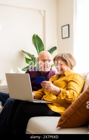 Ältere Ehepaare sitzen zusammen auf einem Sofa und kaufen online mit einem Laptop und einer Kreditkarte. Seniorenpaar sieht fröhlich aus, während sie online mit Shooping Stockfoto