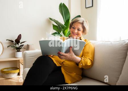 Lächelnde Seniorin, die in einem Wohnzimmer sitzt und ein Buch liest. Eine Seniorin verbringt seine Freizeit damit, ein Buch zu Hause zu lesen. Stockfoto