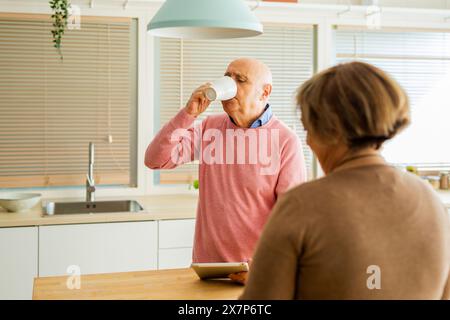 Seniorenmann trinkt Kaffee aus einer weißen Tasse, während eine Seniorin, die eine Tablette hält, vor ihm steht. Stockfoto