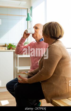 Senior Mann trinkt aus einem weißen Becher und eine Senior Frau hält eine Schüssel. Seniorenpaar, das in der Küche sitzt und einen ruhigen Moment miteinander teilt. Stockfoto