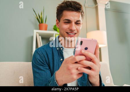 Hübscher junger Kerl, der mit seinem Smartphone im Internet surft, im Wohnzimmer zu Hause auf einem Sofa sitzt. Männer, die online wetten, Nachrichten schreiben oder über soziale Medien mit einer Handy-App teilen. Hochwertige Fotos Stockfoto