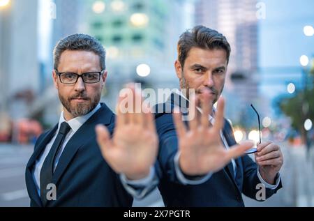 Krise, Risiko. Zwei gutaussehende Geschäftsleute im Anzug hören auf mit der Hand zu singen. Warnausdruck mit negativer und ernsthafter Geste im Gesicht. Halt die Hand an Stockfoto
