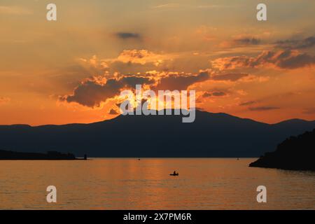 Einsamer alter Fischer, der in einem kleinen Boot in der Bucht vor dem hohen Berg bei Sonnenuntergang mit bewölktem Himmel im Hintergrund für seine Familie fischt Stockfoto