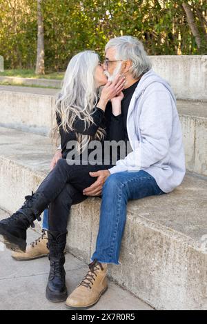 Ein älteres Paar genießt einen zärtlichen Moment, teilt einen Kuss, während es draußen auf einem Betonschritt sitzt, und zeigt dauerhafte Liebe und Gemeinschaft in seinem Leben Stockfoto