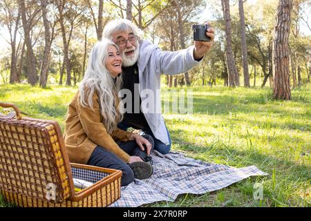 Ein fröhliches Seniorenpaar macht ein Selfie, während es ein sonniges Picknick in einem üppig grünen Park genießt und einen liebevollen Moment ihres Dates zeigt Stockfoto