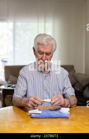 Ein älterer Mann misst sorgfältig seinen Blutzuckerspiegel mit einem Glukometer an einem Tisch in einem gut beleuchteten Wohnzimmer, was einen Moment der persönlichen Heilung darstellt Stockfoto