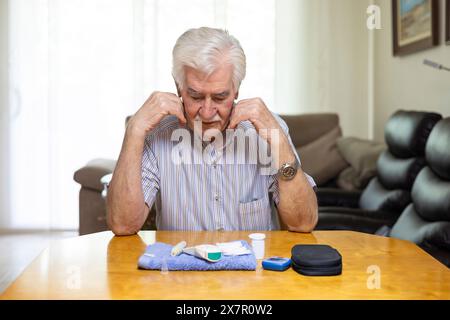 Ein älterer Mann sitzt an einem Tisch, der an Hypoglykämie-Symptomen leidet, während er seinen Blutzuckerspiegel überprüft, mit medizinischen Vorräten in der Nähe Stockfoto