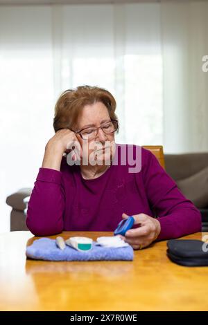 Eine ältere Frau sitzt an einem Tisch und sieht beunruhigt aus, als sie ihren Blutzucker mit einem Glukometer kontrolliert Stockfoto