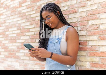 Eine junge Frau genießt einen sonnigen Barcelona-Tag, lässig in Sommerkleidung gekleidet und lächelt, während sie ihr Smartphone vor einer Backsteinmauer benutzt Stockfoto