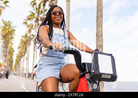 Eine fröhliche Frau genießt eine sonnige Fahrradtour im Sommer auf einem palmengesäumten Weg in Barcelona, die sowohl die Schönheit der Stadt als auch die Freizeitaktivität zeigt Stockfoto