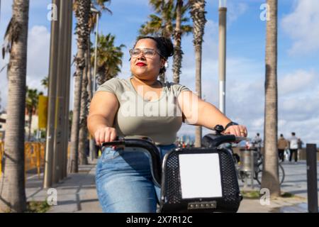 Eine Frau genießt eine sonnige Fahrradtour im Sommer auf einem palmengesäumten Weg in Barcelona, die sowohl die Schönheit der Stadt als auch die Freizeitaktivität zeigt Stockfoto