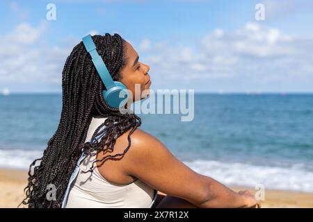 Eine junge Frau genießt Musik mit blauen Kopfhörern, während sie sich an einem sonnigen Sommertag am Strand von Barcelona entspannt Stockfoto