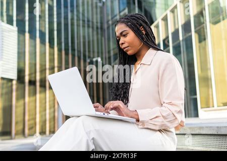 Eine fokussierte professionelle Frau arbeitet an ihrem Laptop außerhalb moderner Gebäude in Barcelona und verkörpert im Sommer eine Mischung aus Arbeit und Freizeit Stockfoto