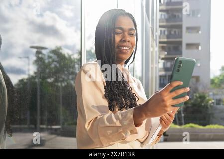 Eine fröhliche Frau mit geflochtenen Haaren genießt einen Sommertag in Barcelona und interagiert mit Freunden über ihr Smartphone Stockfoto