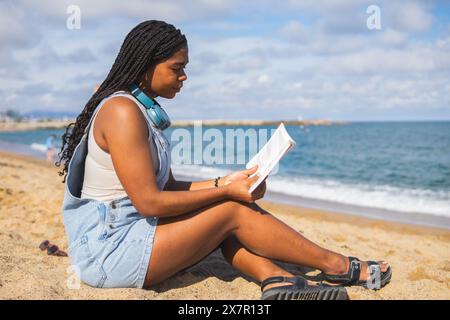 Eine junge Frau genießt die Einsamkeit, indem sie im Sommer an einem Sandstrand in Barcelona ein Buch liest, mit dem Meer im Hintergrund Stockfoto