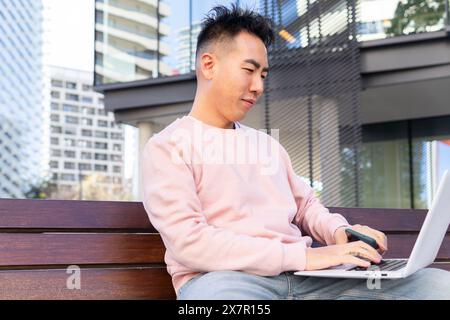 Ein asiatischer Mann in einem lässigen rosa Sweatshirt arbeitet an seinem Laptop, sitzt auf einer Bank mit Wolkenkratzern im Hintergrund. Stockfoto